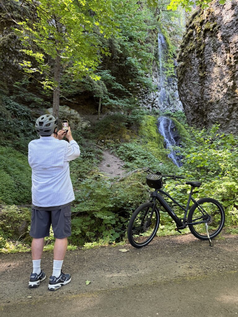 E-bike rider in white shirt standing on the trail admiring a scenic waterfall on the Historic Columbia River Highway near Hood River